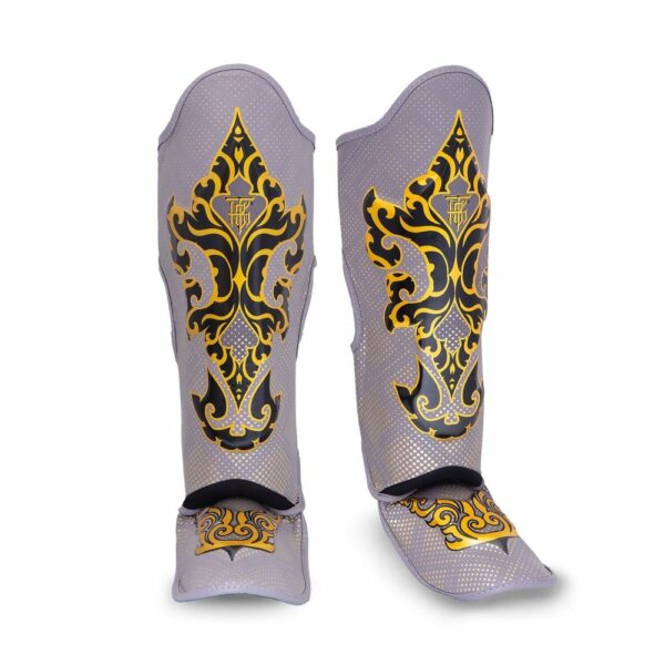 Paire de protège-tibias TopKing modèle Kanok, gris avec motifs dorés inspirés de l’art traditionnel thaïlandais. Design incurvé avec rembourrage et sangles pour un maintien optimal, adaptés aux sports de combat comme le Muay Thai ou le kickboxing.
